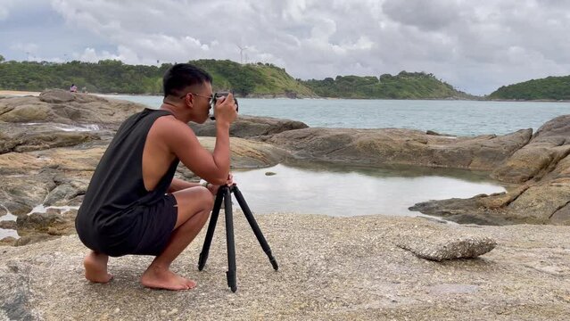 Asian photographer with dslr camera and tripod taking photos by the sea on rocky coast. Filipino young man capturing natural beauty in Phuket, Thailand. Summer tourism, travel destination concepts