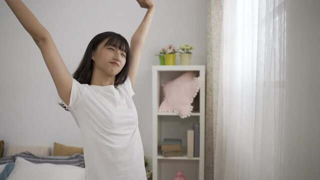waist up of a refreshed asian girl standing near the window and stretching up hands after getting up in the morning at a modern bright home interior