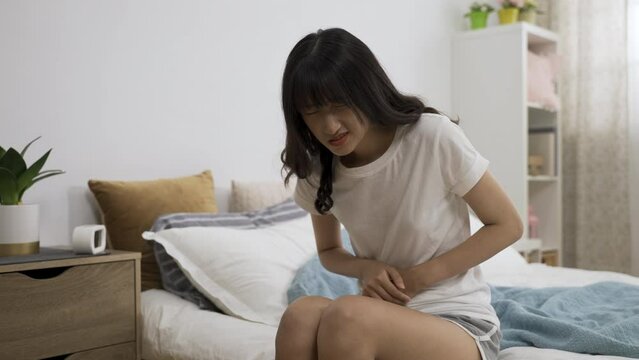 asian young woman sitting on bed pressing her belly with a moan while having pain in her lower abdomen after getting up in the bedroom at home