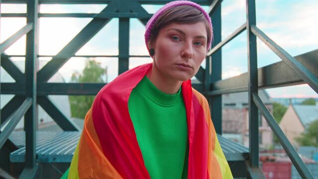 Portrait of an attractive young Caucasian woman wearing LGBT rainbow flag behind back and looking seriously at camera while sitting on stairs. Shot of girl in colorful outfit outdoor. City