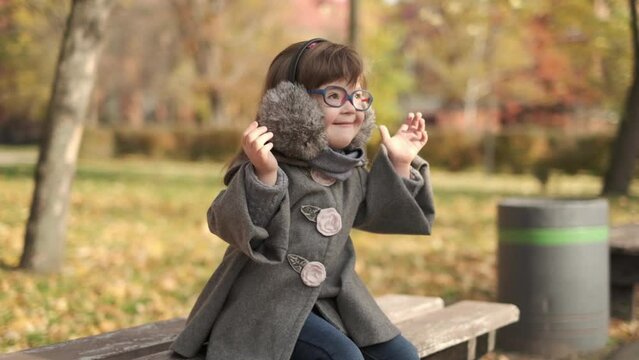 Stylish little girl with brown hair and fashionable eyeglasses wearing warm gray coat and pink boots sitting on bench and holding bouquet of lush foliage, pastime in beautiful warm autumn park