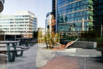 businesswoman reflected on the glass window of the hotel cafe sitting at a small table with tablet and notebook while looking away in the financial center of the city