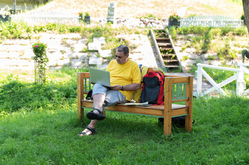 Fototapeta premium Middle-aged man man sitting on the wooden bench in park and using laptop, working outdoors