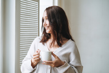 Young beautiful woman forty year with brunette long hair in cozy knitted cardigan with cup of tea...