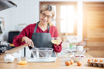Beautiful young woman  is mixing batter, looking at camera and smiling w hile baking in kitchen at...