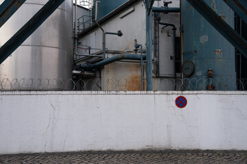 Industrial piping of a chemical plant. Painted in turquoise. Rusty steel construction, barbed wire and a white painted wall with traffic sign 