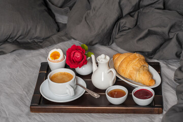 Breakfast in bed with espresso coffee, croissant, honey and jam on the wooden tray, close up. White cup of coffee on the gray bedsheet with pillow and blanket