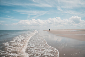 Two old people going for a walk on the beautiful endless sand beach on the Northsea coast of the german island Juist. Some waves of the sea and blue clouded sky. - obrazy, fototapety, plakaty