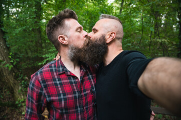 Attractive hipster bearded gay couple taking selfie in the woods. Long beard homosexual men kissing each other outdoor.