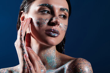 portrait of brunette young woman with glitter on body touching face isolated on blue.
