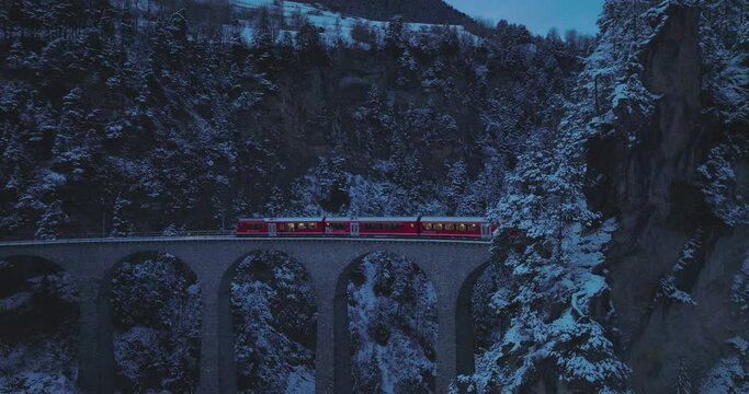 Landwasser Viaduct world heritage sightseeing Glacier and Bernina express in Swiss Alps snow winter scenery at night. Aerial Drone shot red train passing through famous mountain. Filisur, Switzerland.