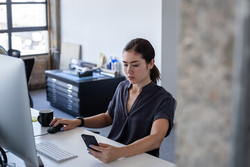 Businesswoman working at a desktop computer in an office looking at a smartphone