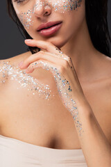 cropped view of young woman with sparkling glitter on cheeks and body touching chin isolated on grey.
