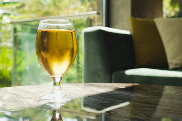 Glass of light filtered beer on the table in front of the sofa. Beautiful sunlight through a beer glass. Place to relax, meeting guests with alcohol soft drinks.