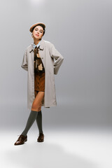 full length of elegant young woman in beret and trench coat posing while looking at camera on grey.