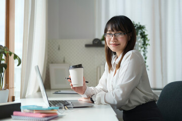 Pretty asian female office worker in eyeglasses holding paper cup of coffee and smiling to camera.