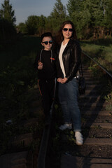 A young mother and son in sunglasses stand holding hands on the railroad tracks against the backdrop of the forest. a family of two