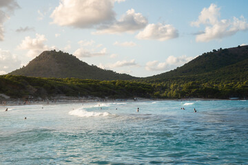 The waves splash on beach of Cala Agulla (Cala Ratjada) on Mallorca in the mediterranean sea. Blue water and a green mountain in the background. People bathing, surfers surfing. Tropical paradise.