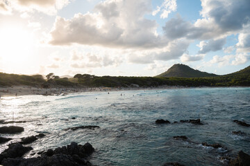 The waves splash on beach of Cala Agulla (Cala Ratjada) on Mallorca in the mediterranean sea. Blue water and a green mountain in the background. People bathing, surfers surfing. Tropical paradise.