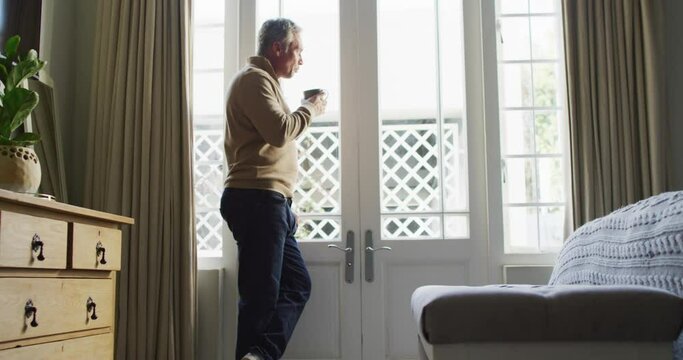 Happy caucasian man looking through window and drinking coffee in bedroom