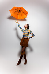 full length of cheerful woman in striped turtleneck and skirt posing with umbrella on grey.