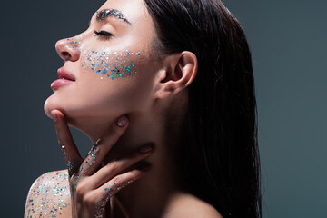 portrait of young brunette woman with closed eyes and glitter on eyebrows touching neck isolated on grey. - obrazy, fototapety, plakaty