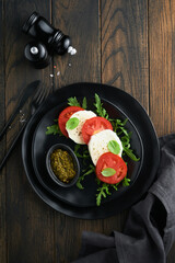 Caprese salad. Italian caprese salad with sliced tomatoes, mozzarella cheese, arugula, basil, olive oil in black plate over old wooden dark background. Delicious Italian food. Top view.