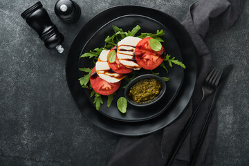 Caprese salad. Italian caprese salad with sliced tomatoes, mozzarella cheese, arugula, basil, olive oil in black plate over old brick tiles black background. Delicious Italian food. Top view.