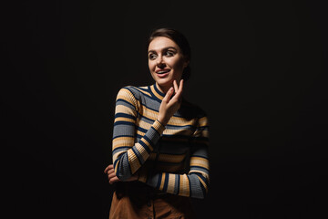 cheerful young woman in striped turtleneck and skirt looking away isolated on black.