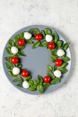 Caprese salad in the form of a Christmas wreath. Festive tomato mozzarella and basil appetizer on grey plate.