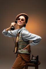 happy young woman in beret and sunglasses holding vintage telephone on brown.