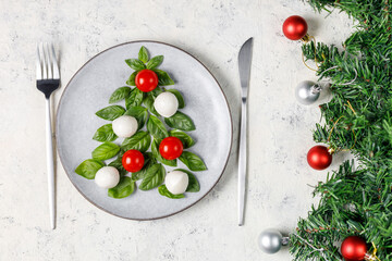 Caprese salad in the form of a Christmas tree. Festive tomato mozzarella and basil appetizer on light plate. - obrazy, fototapety, plakaty