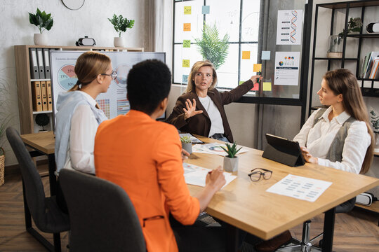 Senior blond financier woman pointing on board with paper sticks during speech on conference with multiracial female colleagues. Competent economists discussing company incomes at office room.