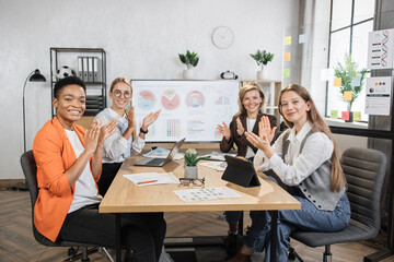 Multi ethnic female partners in formal clothes applauding together while sitting at office. Competent financiers businesswomen with modern gadgets on table looking and smiling on camera.