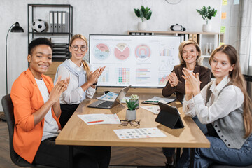 Multi ethnic female partners in formal clothes applauding together while sitting at office. Competent financiers businesswomen with modern gadgets on table looking and smiling on camera.