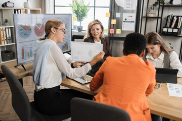 Four multi ethnic female economists sitting at office with various gadgets and analysing financial growth in worldwide economy. Female partners having business meeting.