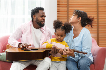 Cute little girl with mom and dad playing guitar and smiling while sitting on the sofa at home....