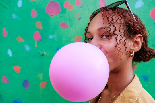 Woman With Curly Hair Blowing Bubble Gum In Front Of Wall