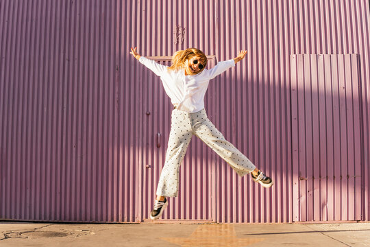 Cheerful woman jumping with arms outstretched in front of corrugated wall