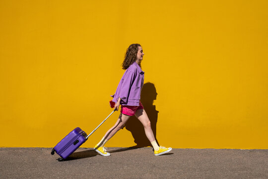 Young woman pulling luggage walking by yellow wall