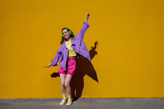 Carefree Woman With Hand Raised Dancing And Listening To Music In Front Of Yellow Wall