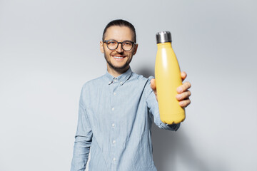 Studio portrait of young smiling man showing close to camera a steel thermo water bottle of yellow color on light grey background.