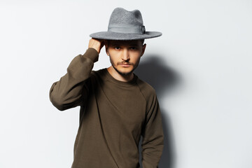 Studio portrait of young man holding wireless earbuds and charger case in hands on white background, wearing sweater and grey hat.