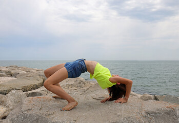 girl performs gymnastic exercises arching her back backwards on the rocks by the sea without...
