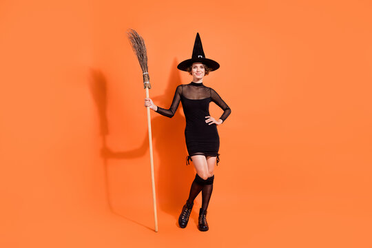 Full size photo of stunning young woman hold broomstick wear stylish black halloween witch outfit isolated on orange color background