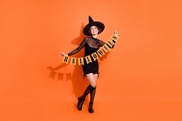 Full length photo of adorable young lady hold garland flags wear stylish black halloween witch clothes isolated on orange color background