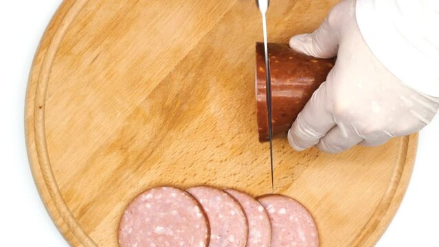 Sausage cutting. A cook in white gloves cuts a red salami sausage into pieces with a large sharp knife. Beef sausage for sandwiches. Pink pork sausage with pieces of lard. Top view. Close-up. Isolated