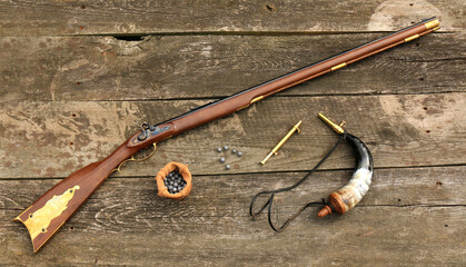 Traditional Kentucky rifle and shooting equipment such as a gunpowder horn, a gunpowder measure and bullets on an old, damaged board.