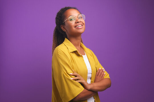 Young Confident Positive African American Woman Proudly Posing With Arms Crossed In Front Of Chest And Raising Chin Looks At Screen Dressed In Yellow Shirt Stands On Purple Studio Background