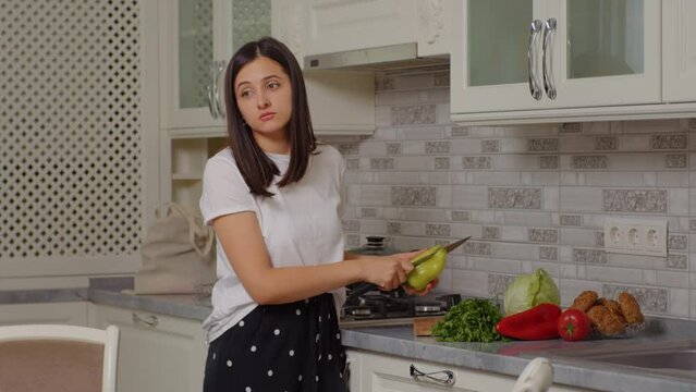 Brunette cutting a green pepper and talking to somebody in the kichen. Homemade dinner with vegetables.